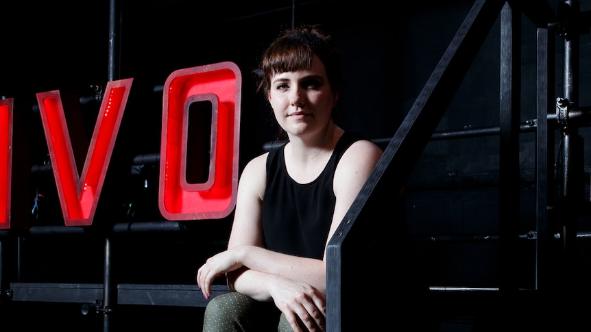 Young woman in black tank top and army-green pants sitting on set with large light-up letters spelling TIVOLI behind her in red.
