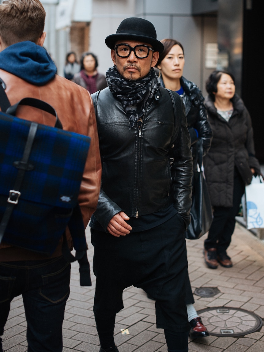 A man in Tokyo wears all black pants, leather jacket, hat and bold eyeglasses.