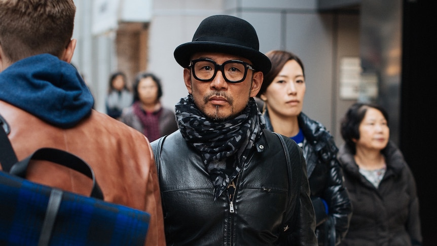 A man in Tokyo wears all black pants, leather jacket, hat and bold eyeglasses.