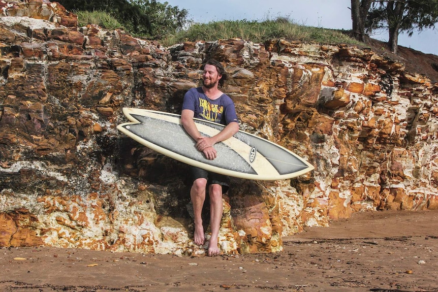 man with a surf board smiling and leaning against rocks