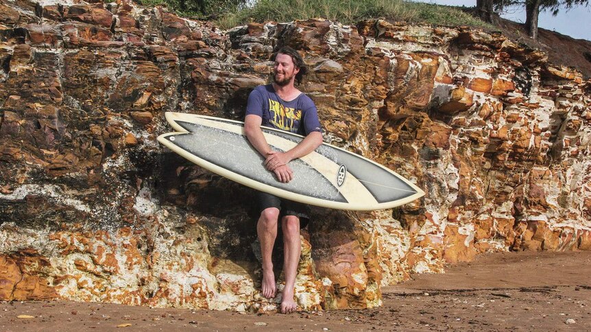 man with a surf board smiling and leaning against rocks