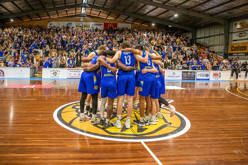 A bunch of basketball players huddle in the middle of a basketball court in front of a packed crowd.