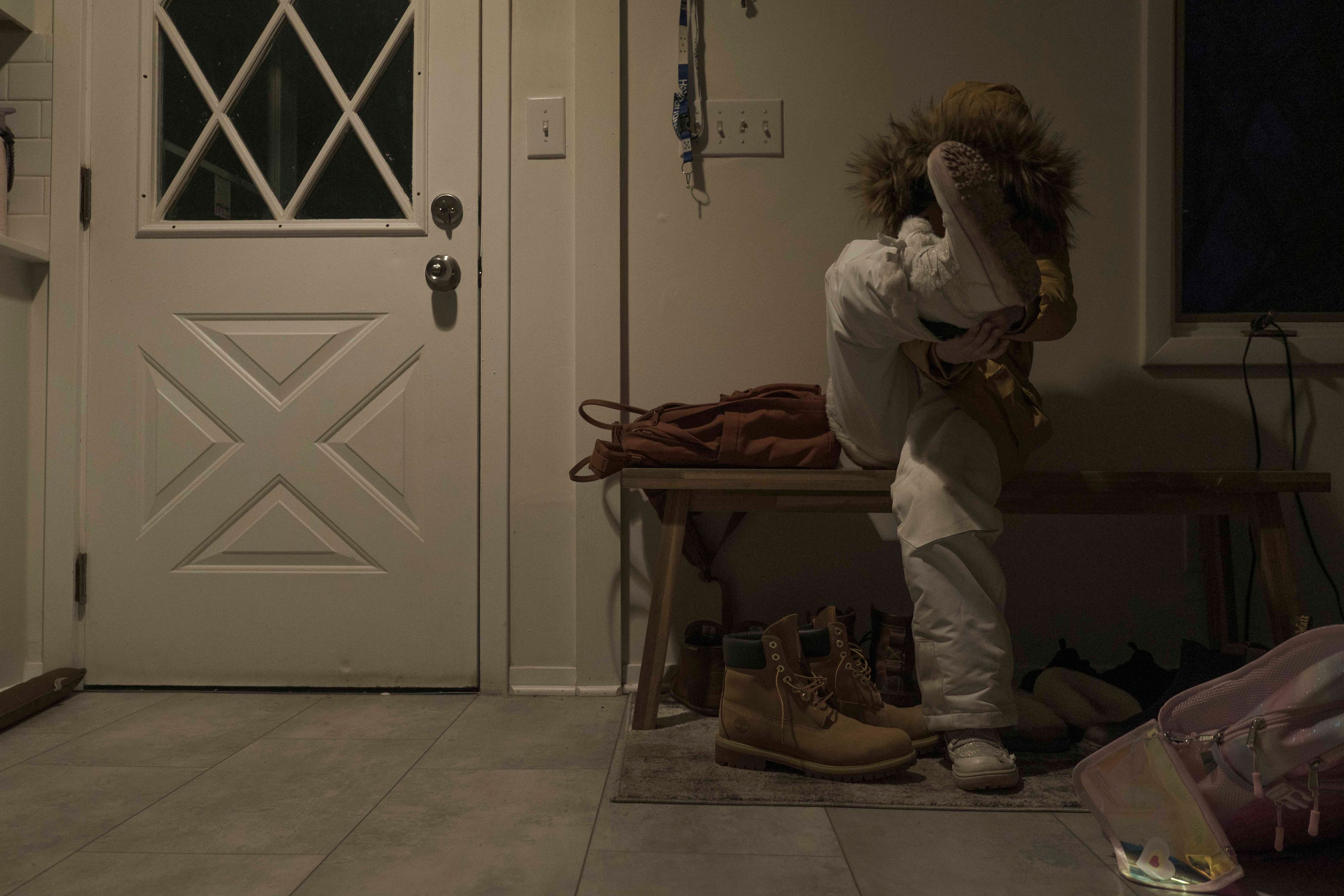 A young child puts on heavy shoes. She is seated by a white door.