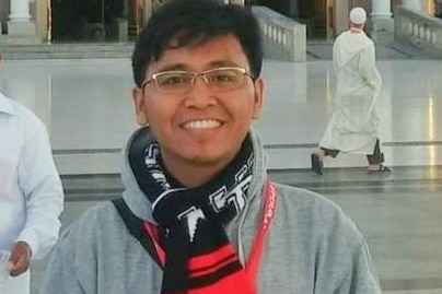 A person with black hair and wearing glasses smiles with a scarf in front of a building.