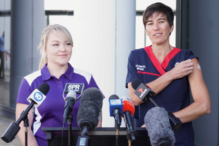Two nurses standing at a podium speaking to journalists, smiling, with one woman showing a bandaid on her arm from the vaccine.