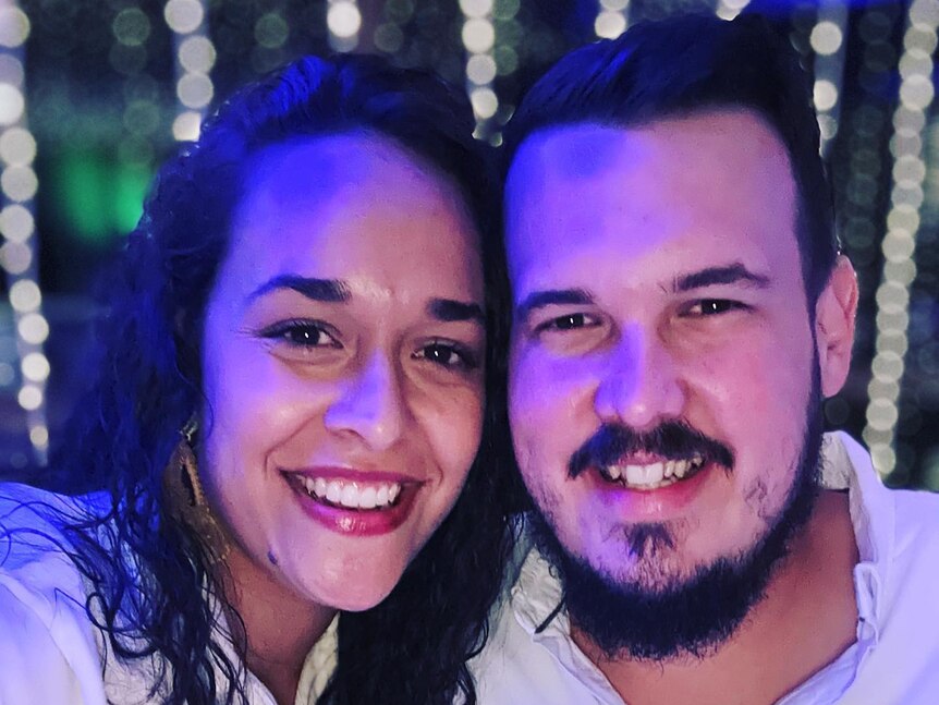 A close up of a couple in their twenties. Their faces are bathed in blue light. They are smiling. 
