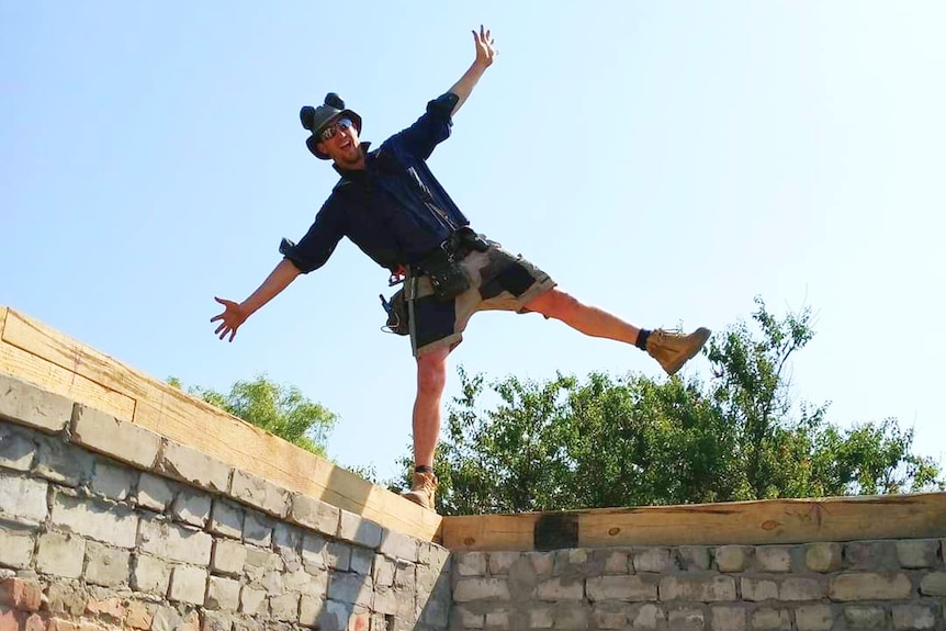 A tradesman with his arms in the air balances on a brick wall