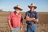 Two smiling older men wearing hats stand in a sunny paddock.
