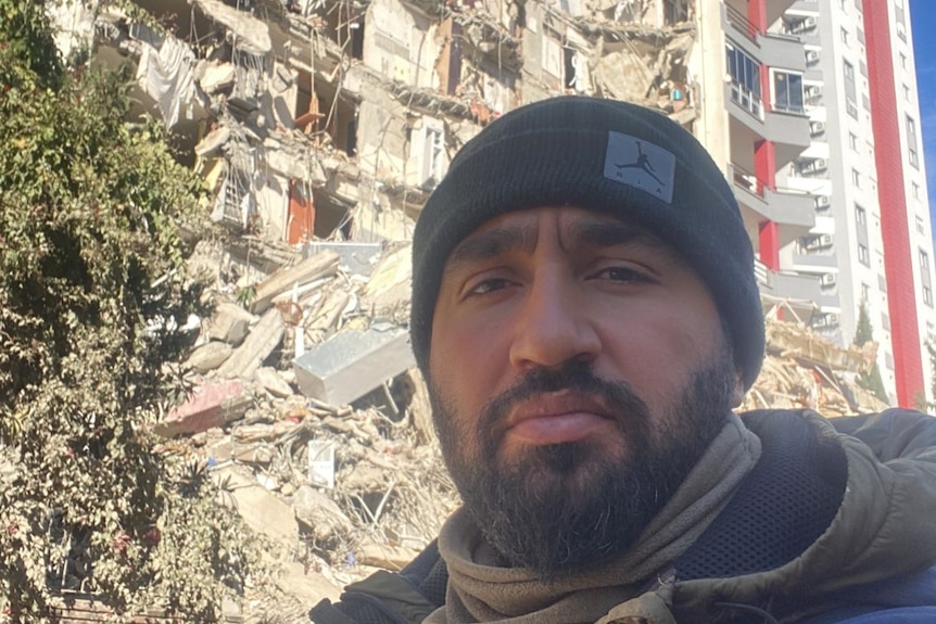 A man with a serious expression stands in front of a damaged building.
