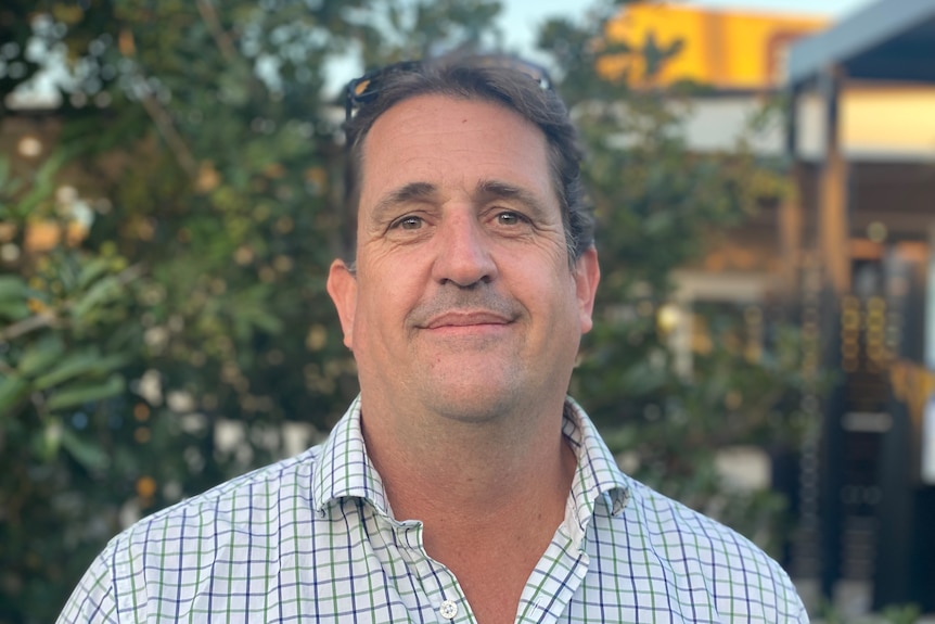 a man stands in a check shirt with trees behind