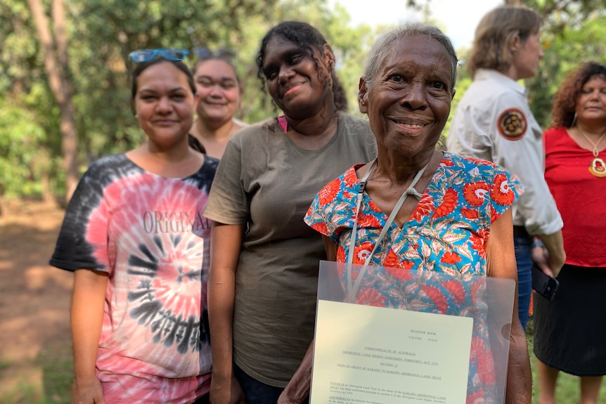 Three female traditional owners standing and smiling while one holds a file full of papers. There's greenery in the background.