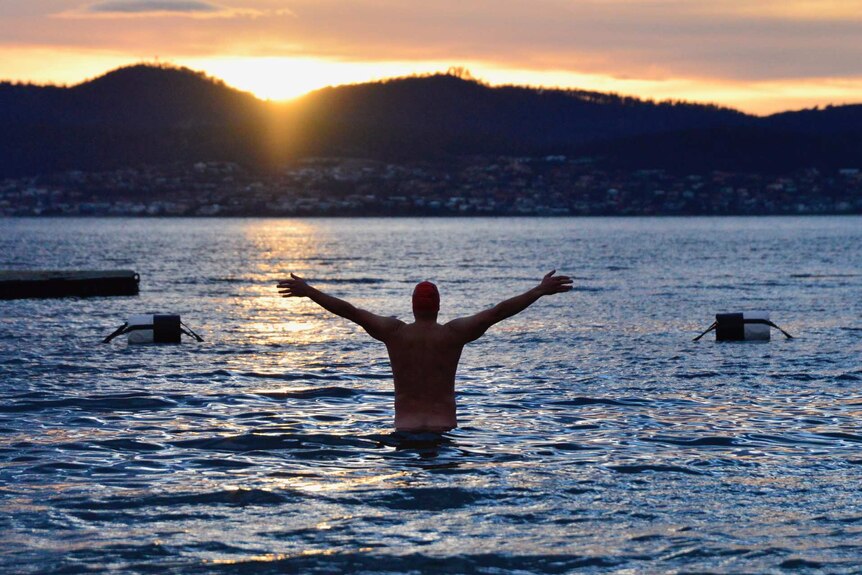 Man raises arms in water as sun rises.