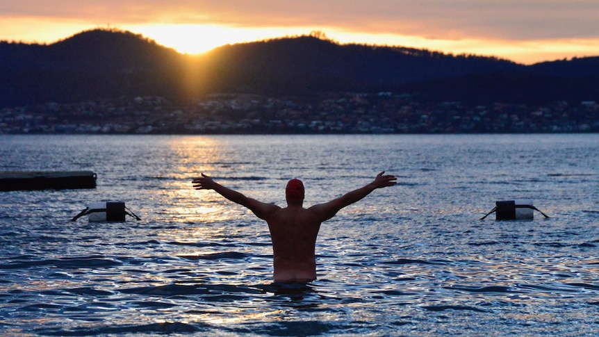 Man raises arms in water as sun rises.