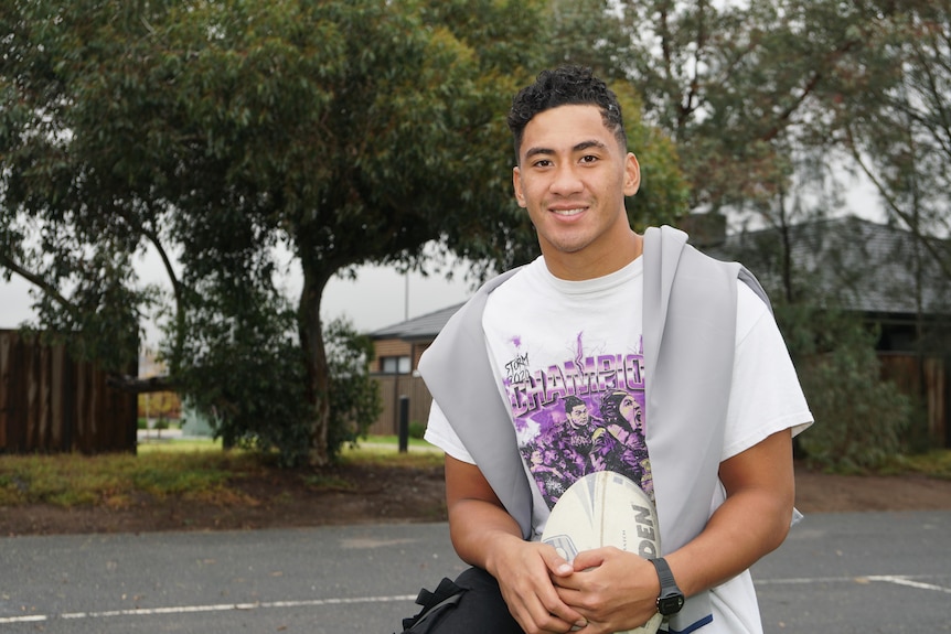 A young man in a Melbourne Storm t-shirt hugs a rugby ball in a carpark.