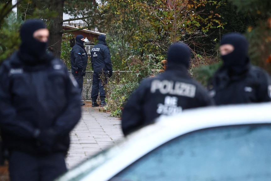 Police in tactical gear and balaclavas stand around on a footpath. 