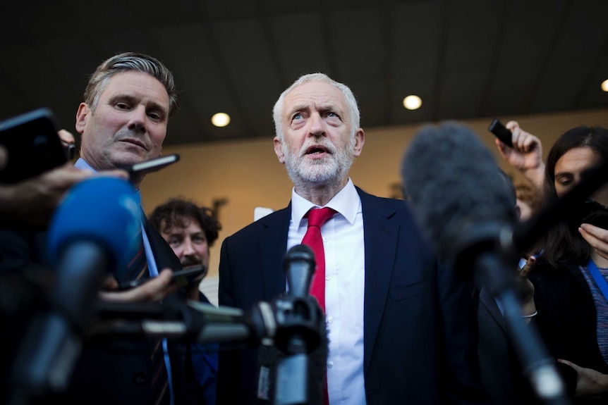 A man with a beard stands surrounded by reporters and microphones