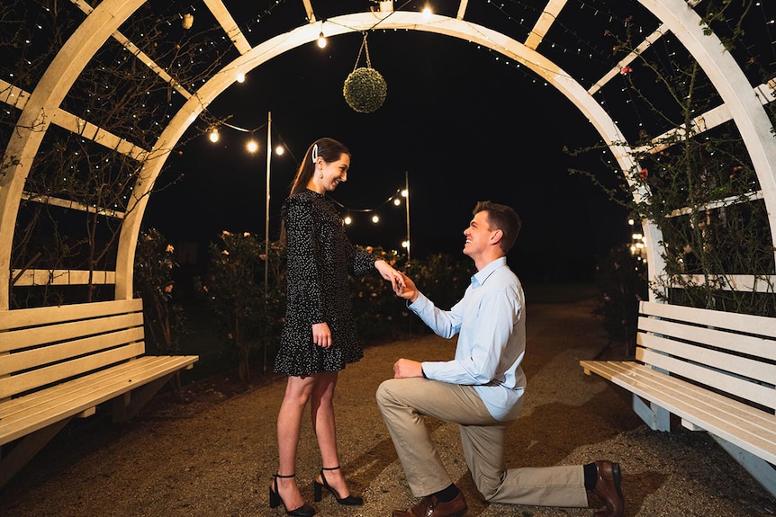 Adam Ison proposes on bended knee to Brielle Musgrove under an archway at night with fairy lights.
