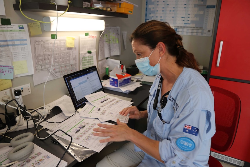 A health worker wearing a facemask sits at an office desk and sorts through paperwork.