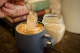 A teaspoon of sugar is poured into a cup of coffee on a wooden table.