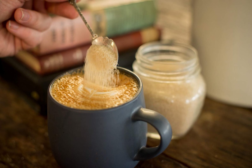 A teaspoon of sugar is poured into a cup of coffee on a wooden table.
