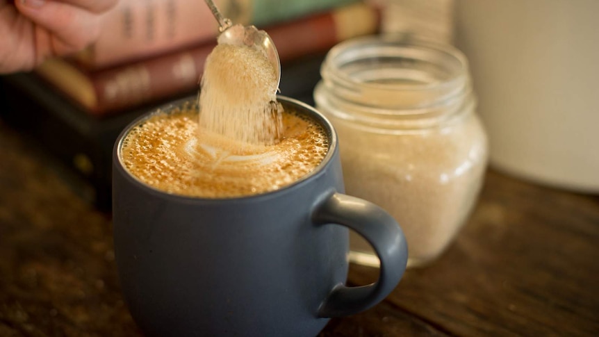 A teaspoon of sugar is poured into a cup of coffee on a wooden table.