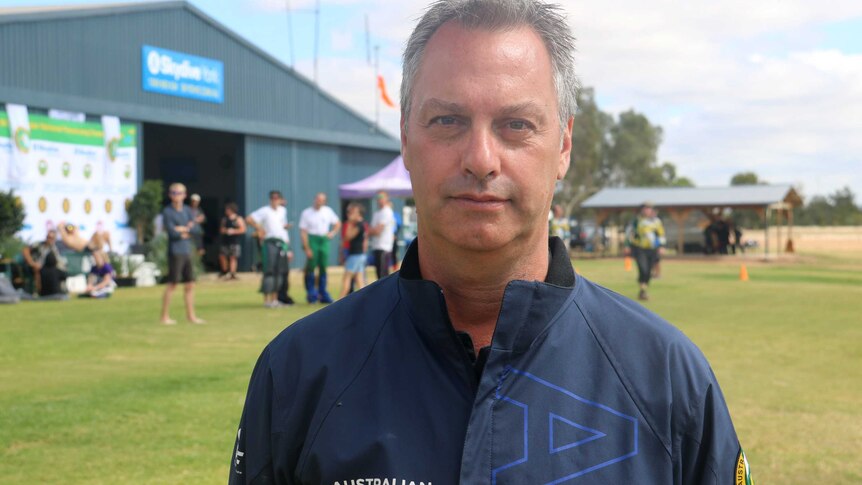 A mid shot of a man in a blue shirt standing in a field looking at the camera with others out of focus in the background.