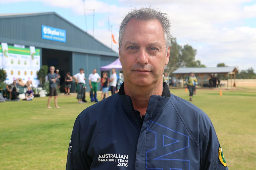 A mid shot of a man in a blue shirt standing in a field looking at the camera with others out of focus in the background.