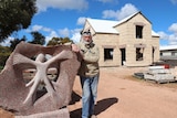 Man in front of stone house leaning on carved stone sculpture.