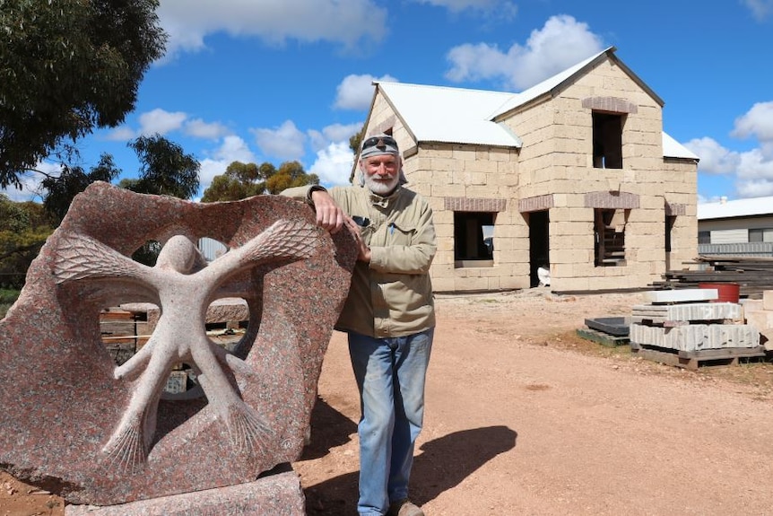 Man in front of stone house leaning on carved stone sculpture.