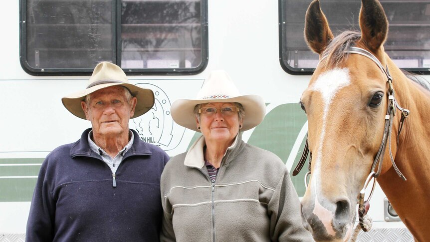 A man and a woman standing in front of a caravan holding one of their horses.