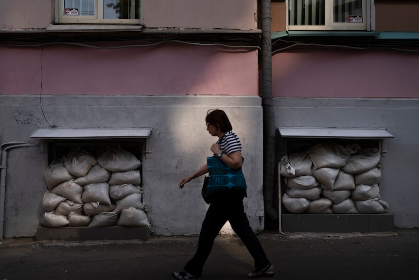 A woman carrying a bag is walking past an apartment building with sandbags stacked against windows