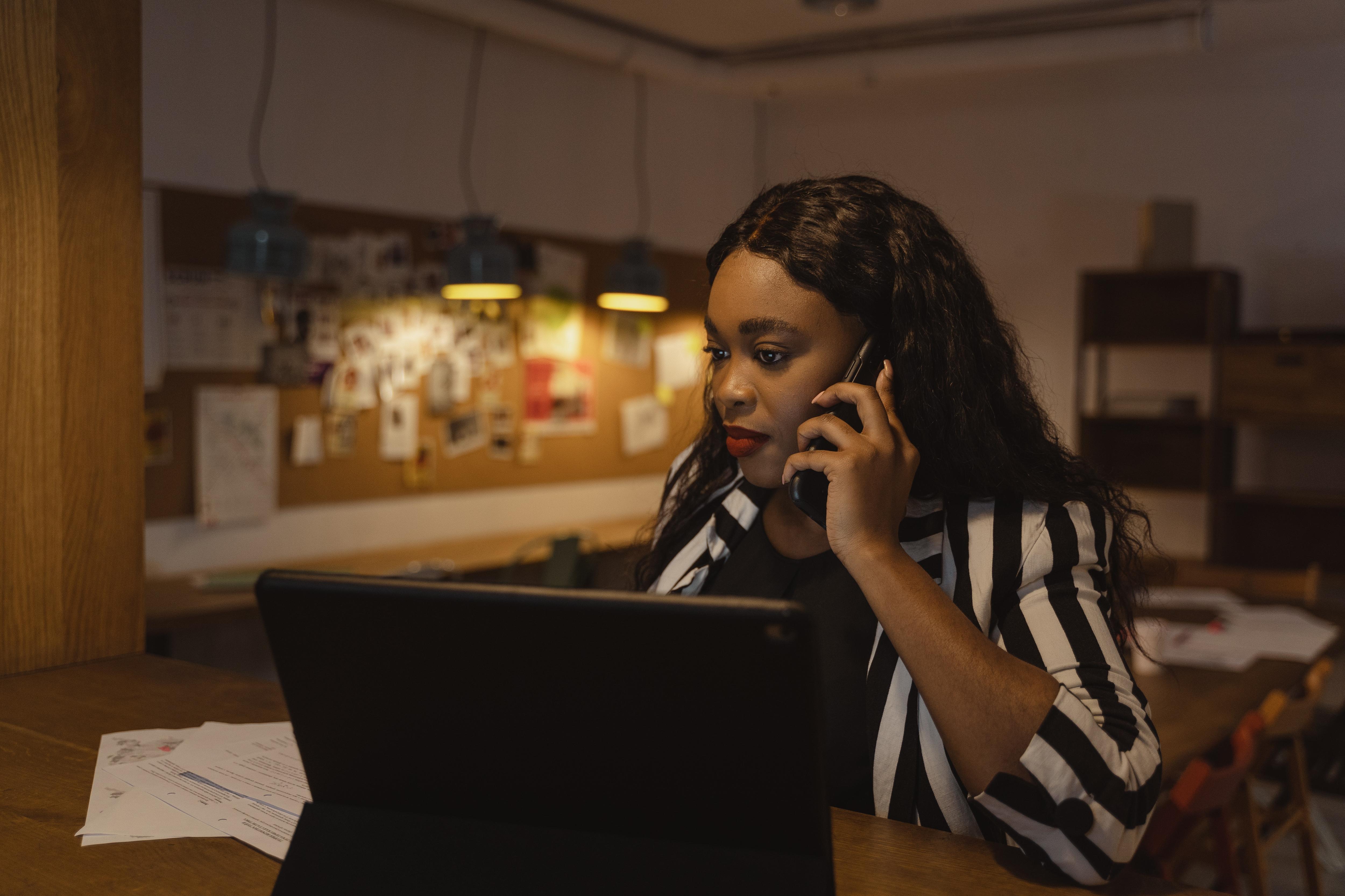 A woman in a black and white shirt at a kitchen table at home with her computer open talking on the phone