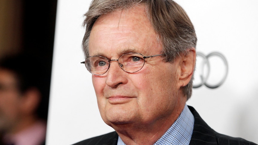 An older white man in a suit and glasses smiles in front of a white photo backdrop.