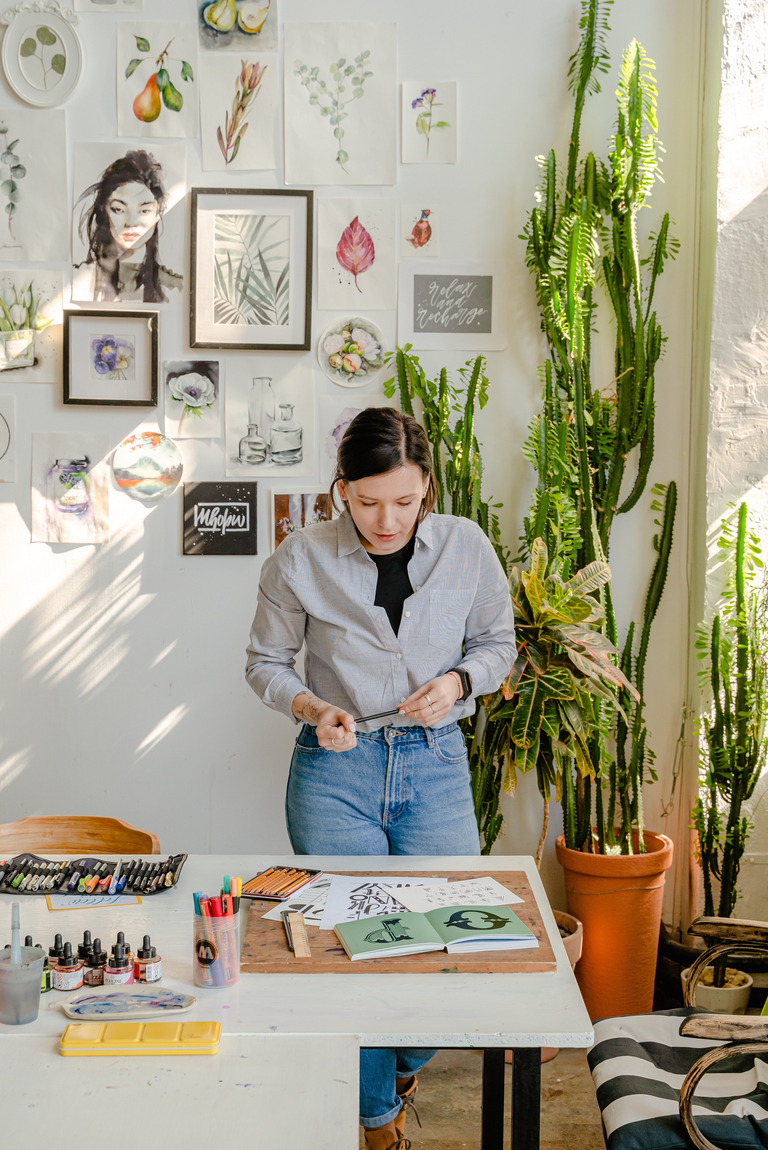 Woman stands at a table looking at artwork against a backdrop of pictures and plants