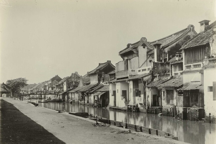 A black and white image shows a canal lined with Dutch colonial-era terraces.