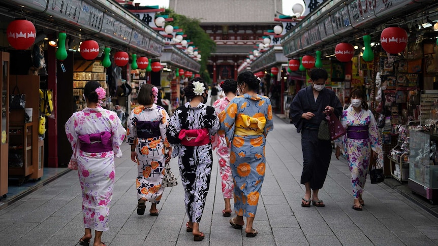 A group of women in Japanese traditional wear walk past two women in face masks in an ornate temple.