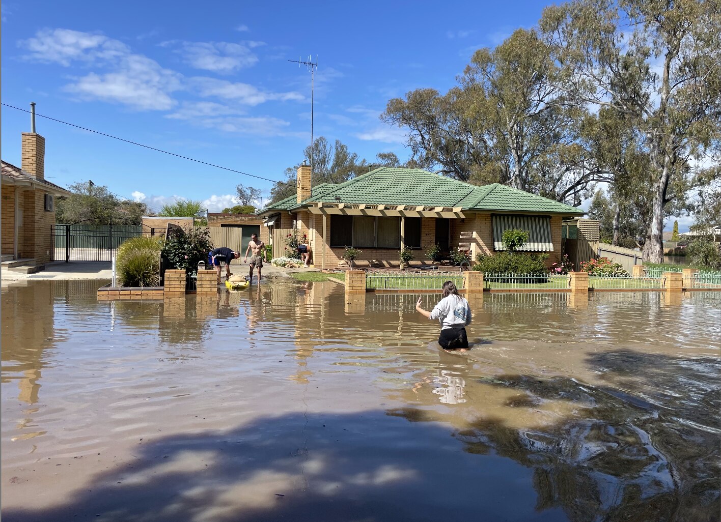 A lady wades through thigh-high water on the street as several people help to sandbag the front of a house. 
