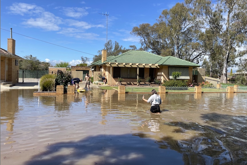 A lady wades through thigh-high water on the street as several people help to sandbag the front of a house. 