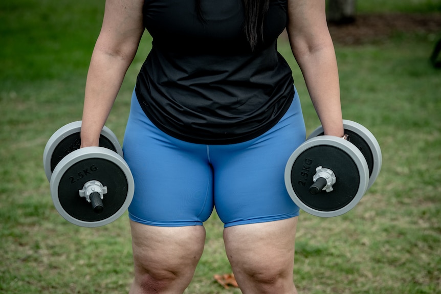 A close-up of Xanthia's disroportionately larger lower body as she holds two large handweights by her side.