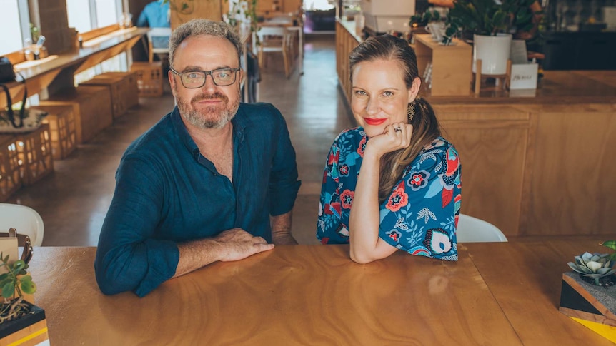 A man with curly grey hair and a beard, and a woman with long, dark blonde hair, sitting in a cafe.