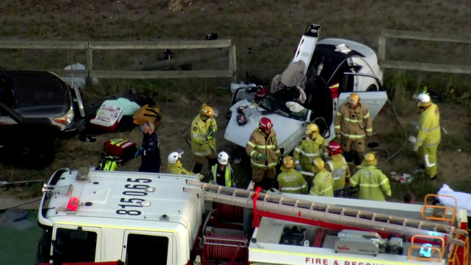 An aerial shot showing a crowd of firefighters and a fire truck next to the wreckage of a white car after a road crash.