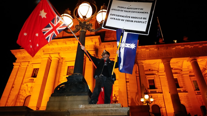 a man wearing dark clothing and a mask waves the Australian red ensign and Eureka flags on the steps of Parliament house at nigh