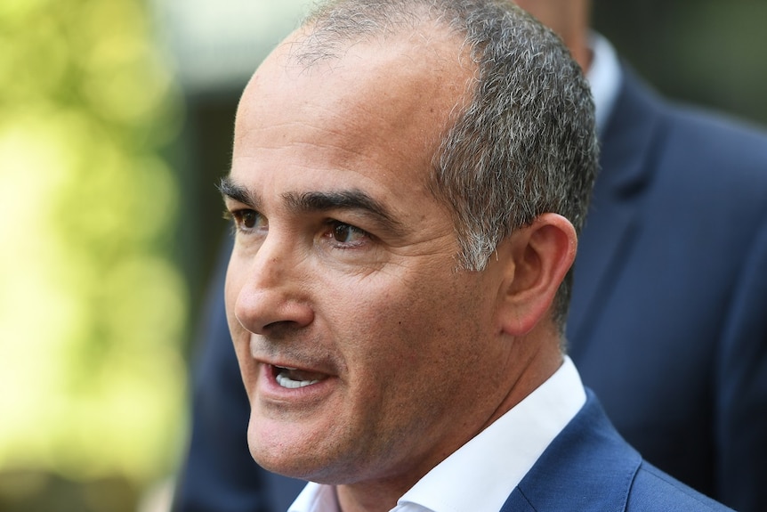 James Merlino, dressed in a white shirt and blue jacket, delivers a press conference outdoors.