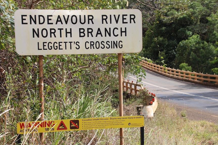 A sign for Leggett's Crossing stands on the side of a road in front of a bridge.