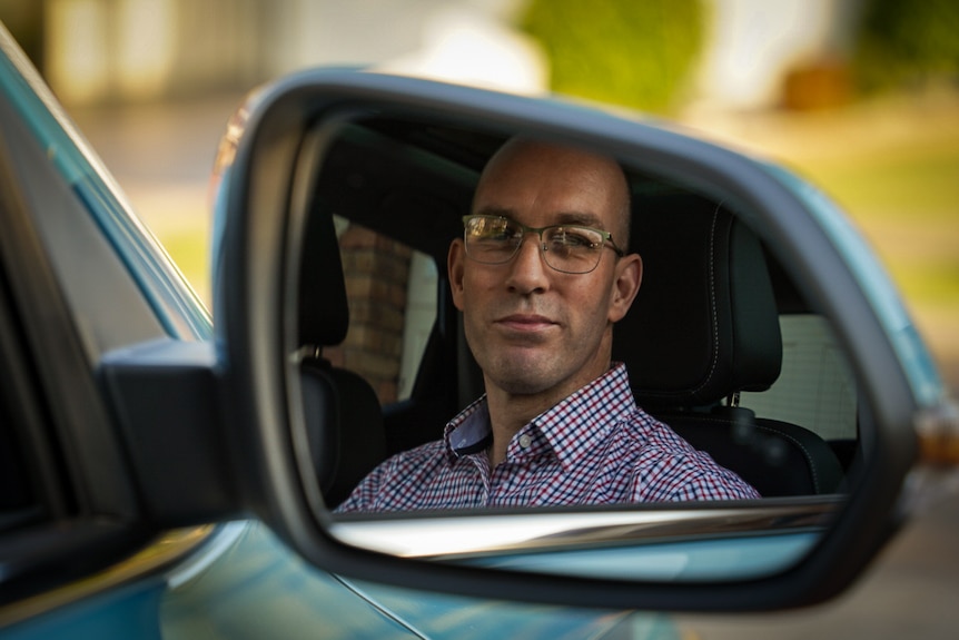 The side mirror reflection of a man with glasses sitting in a blue car 