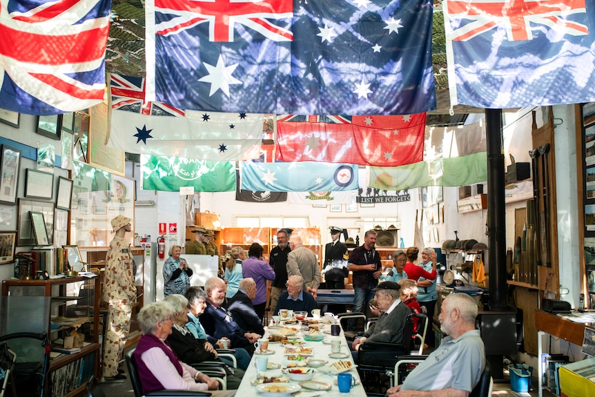 Older folks chat around a long table filled with tea, sandwiches and treats in a room with flags hanging from the ceiling.
