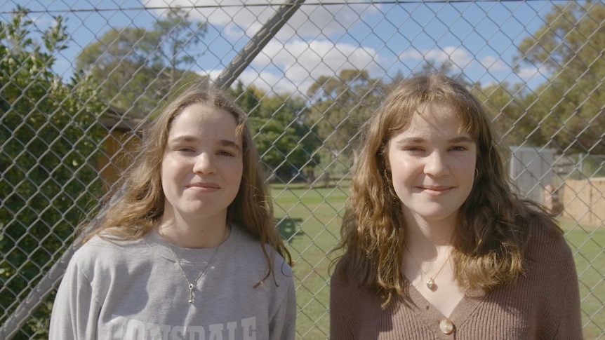 Lucy and Steph face the camera in front of a cyclone wire fence.
