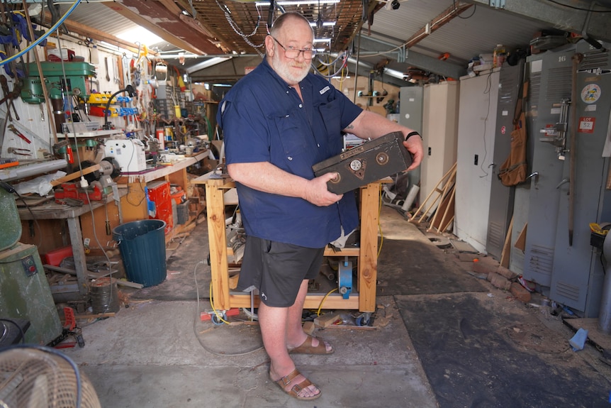 A man wearing a blue shirt and shorts holds a metal box in a shed