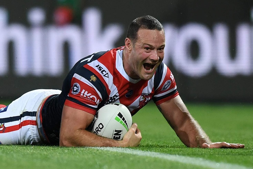 A male NRL player smiles as he slides on grass holding a football while scoring a try.
