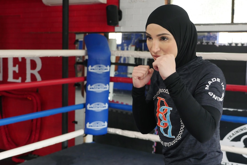 A female boxer wearing a hijab is in the boxing ring, bare hands in front of her face.